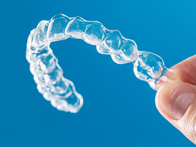 The image shows a clear plastic dental retainer with multiple ridges, held up by a hand against a blue background.