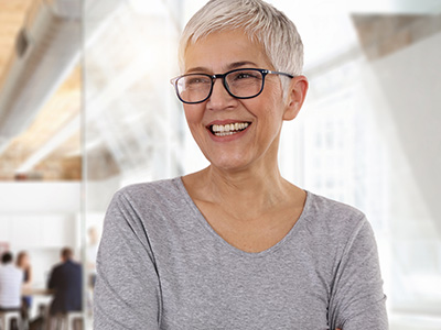 A woman with short, gray hair and glasses is smiling while standing in an office environment.