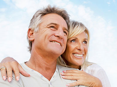 This image features an older couple, a man and woman, embracing each other with smiles on their faces. They are outdoors during the daytime under clear skies.
