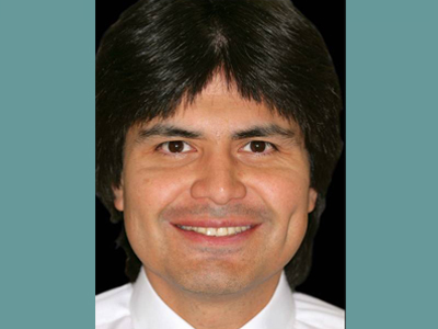 The image is a portrait of a man with dark hair, wearing a white shirt and a tie. He has a smile on his face and appears to be looking directly at the camera.