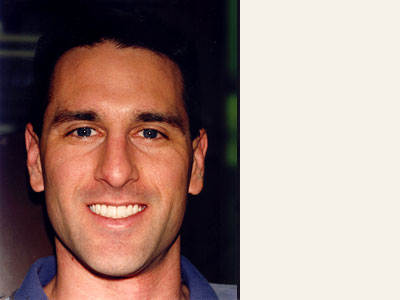 The image is a photograph of a man with short hair, wearing a blue shirt and smiling. He appears to be indoors, possibly in an office setting.