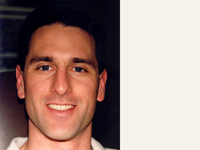 The image shows a man with short hair, smiling slightly and looking directly at the camera. He is wearing a light-colored shirt.