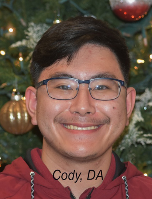 The image shows a person smiling at the camera, wearing glasses and a red top. They are standing in front of a Christmas tree with decorations.