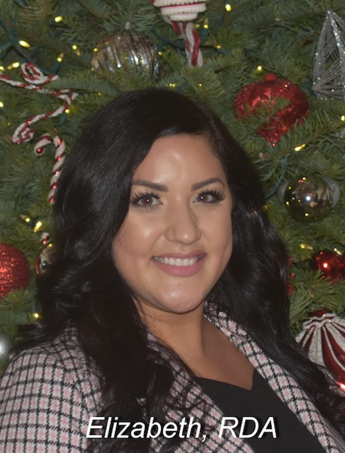 The image shows a woman posing for a portrait in front of a Christmas tree, with festive decorations and lights visible behind her. She is smiling and wearing a checkered blazer over a black top, with her hair styled down.