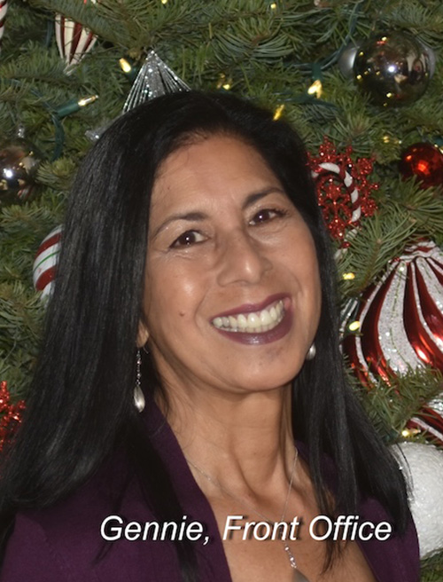 A woman with dark hair and a purple lipstick smile, wearing a black top, stands in front of a decorated Christmas tree.