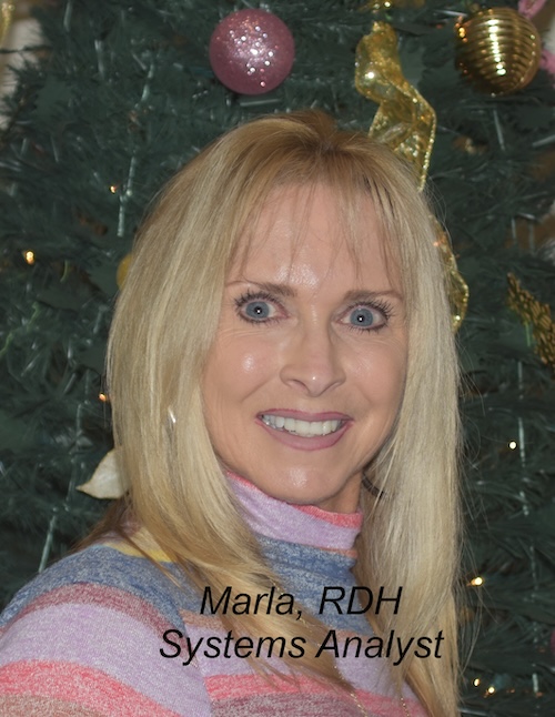 The image shows a woman standing in front of a Christmas tree, posing for the photograph with a smile. She has blonde hair and is wearing a colorful top.