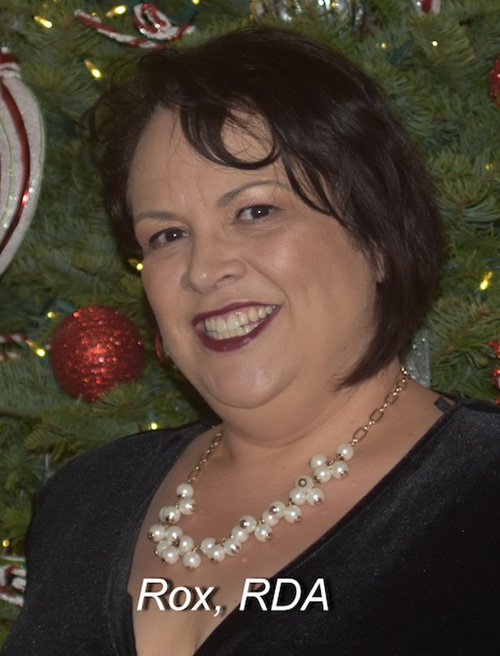 The image is a photograph of a person standing in front of a decorated Christmas tree. The individual appears to be an adult woman with short hair, wearing makeup and a dark top with a necklace. She has a slight smile and is posing for the photo. In the foreground, there is text that reads  Rox, RDA.