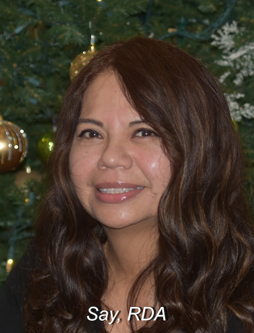 The image shows a woman standing in front of a Christmas tree, smiling at the camera. She has dark hair and is wearing a black top with a light-colored shirt underneath. The text on the image reads  SAY, RDA.