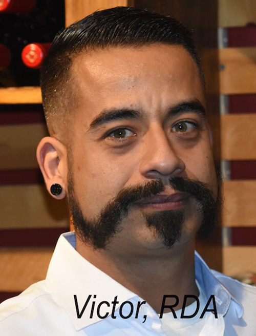 The image is a photograph of a man with a beard and mustache, wearing a white shirt and black sunglasses. He has tattoos on his arms and a visible earring. In the background, there are bottles and wine glasses, suggesting a social or dining setting.