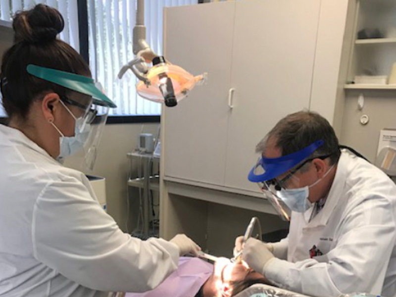 Two people in a medical setting, one wearing protective eyewear and the other using dental tools on a patient s mouth.
