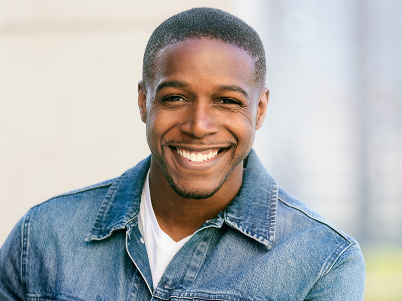 The image features a smiling man with short hair, wearing a denim jacket and standing against a neutral background.