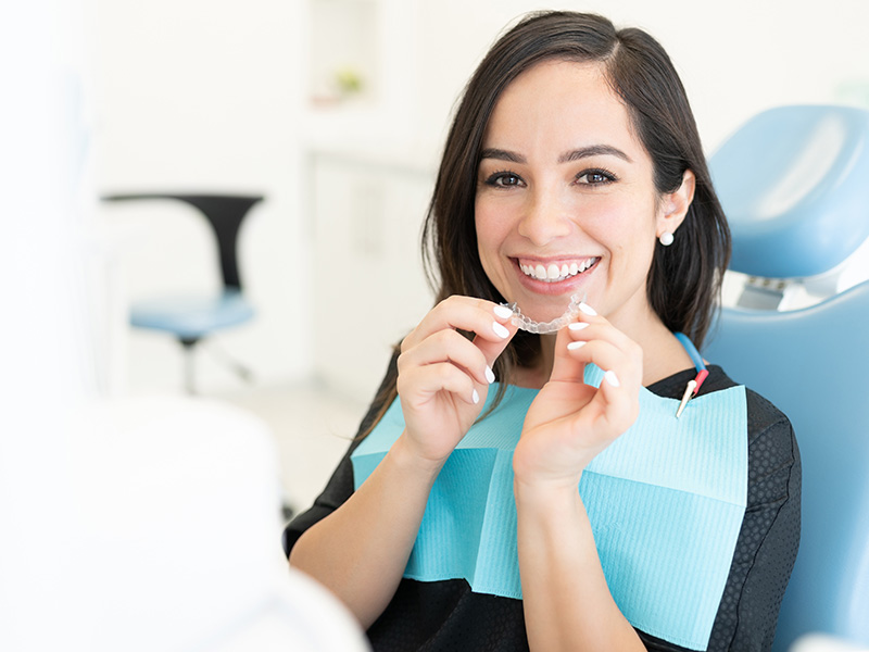 The image depicts a dental hygienist in a professional setting, smiling at the camera while holding a toothbrush and a dental mirror.