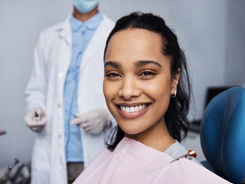 A woman with a bright smile, sitting in a dental chair, receiving dental care from a professional wearing protective gloves and a mask.