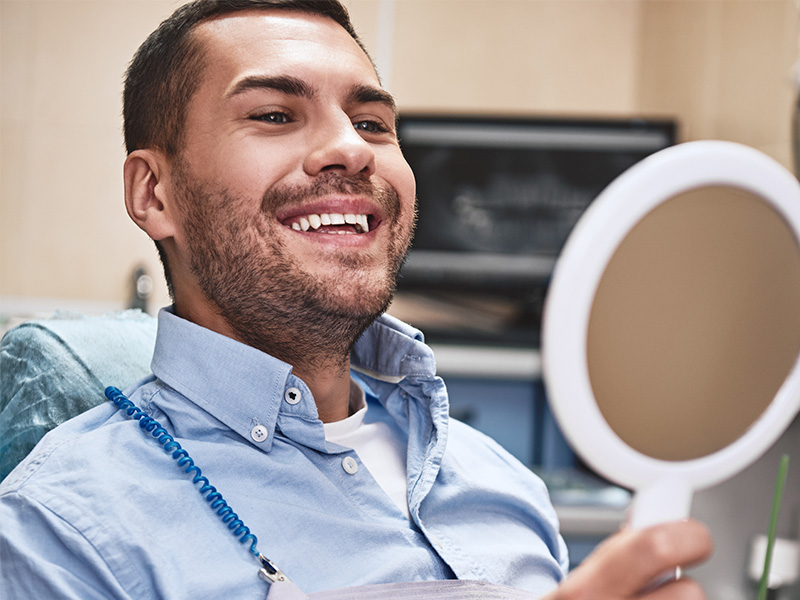A man is smiling at his own reflection in a mirror, sitting in a barber chair with a necklace around his neck.
