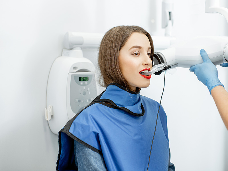 In the image, a woman is seated in a dental chair, receiving treatment from a dentist who is using a device with a clear tube attached to it. The woman appears to be relaxed and engaged in the process, while the dentist focuses on her oral health care.