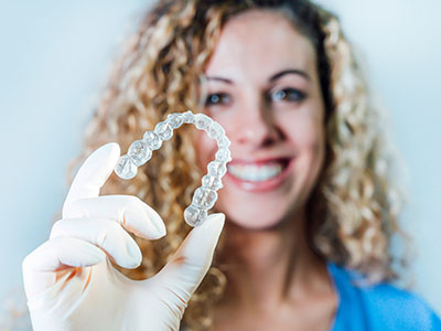 A woman in a blue shirt is holding a transparent dental retainer with her right hand, smiling at the camera.