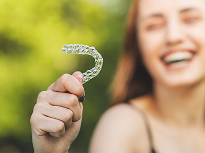 An image of a person holding up a large, clear dental retainer with a smile, set against a blurred outdoor background.