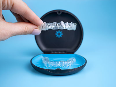 An image of a person s hand holding an open box containing a set of clear dental aligners, with the aligners placed on a blue tray.