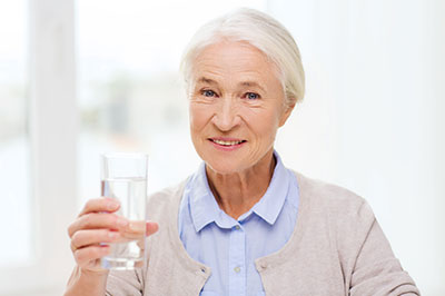 An elderly woman is smiling and holding a glass of water.