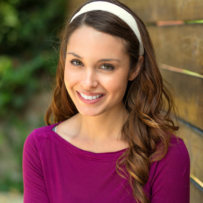 Smiling woman with long brown hair, wearing a purple top and headband, standing in front of a wooden fence.