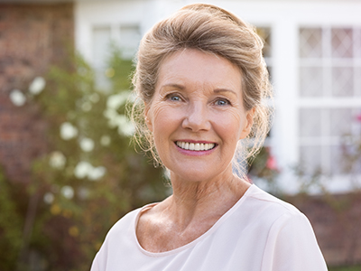 A smiling older woman with gray hair, wearing a white top and standing in front of a house with a brick wall and a window.