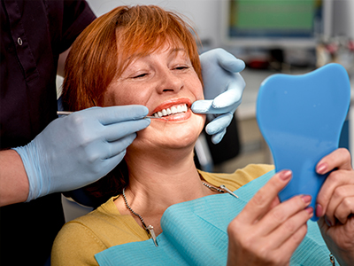 A woman in a dental chair receiving dental care, with a dentist performing a procedure and holding up a blue model of a mouth.