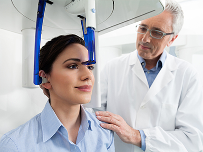 The image shows a woman seated in front of a large, blue, head-mounted device, which appears to be a medical or diagnostic machine. To her right stands a man wearing a white lab coat and glasses, who seems to be examining the device or assisting with its operation.