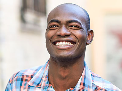 The image features a man with a broad smile, looking directly at the camera. He has short hair and is wearing a plaid shirt. The background is blurred but appears to be an outdoor setting with a building and some foliage.