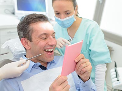 The image shows a person sitting in a dental chair, holding up a pink card or sign with a smile, while a dentist is standing behind them, looking at the card with a slight smile.