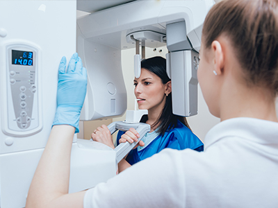 The image shows a woman in a blue lab coat standing next to a large, white CT scanner machine.