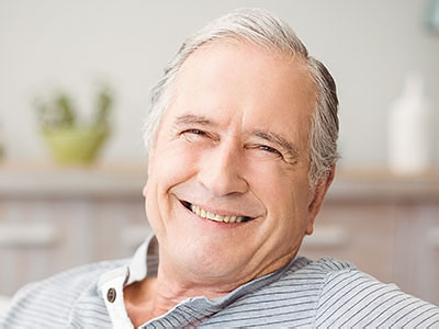 The image shows an older man with gray hair, smiling broadly and looking directly at the camera. He has a light blue shirt and is sitting in what appears to be a comfortable indoor setting, possibly a living room, with a blurred background that suggests a home environment.