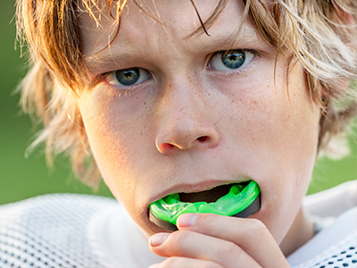 A young boy with blonde hair and a sports jersey is shown in a close-up, brushing his teeth while holding a green toothbrush.