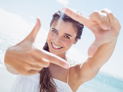 The image shows a woman holding up a peace sign with one hand while the other hand is extended towards the camera, smiling and looking directly at it. She has long hair, fair skin, and is wearing a white top. The background is a bright, sunny outdoor setting with blue sky and white clouds visible.