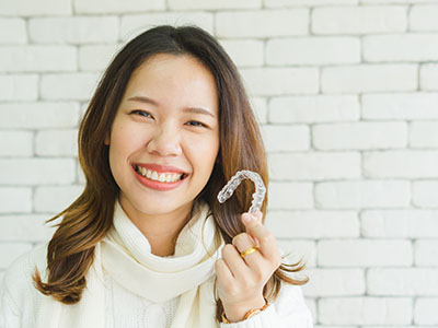 The image features a smiling woman holding up an object that resembles a dental retainer, with a brick wall and white tiles in the background.